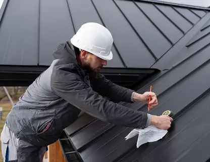 Construction worker in a white helmet measuring a black metal roof with a tape and pencil.